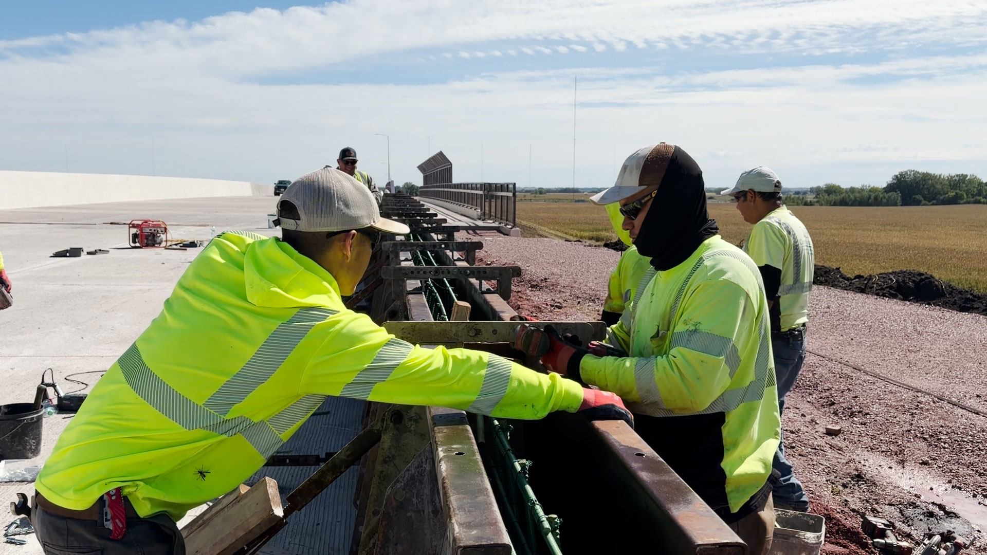 Construction crew members in high-visibility safety clothing install an exterior barrier rail near the BNSF overpass. Work equipment and open farmland are visible alongside the road under a partly cloudy sky.