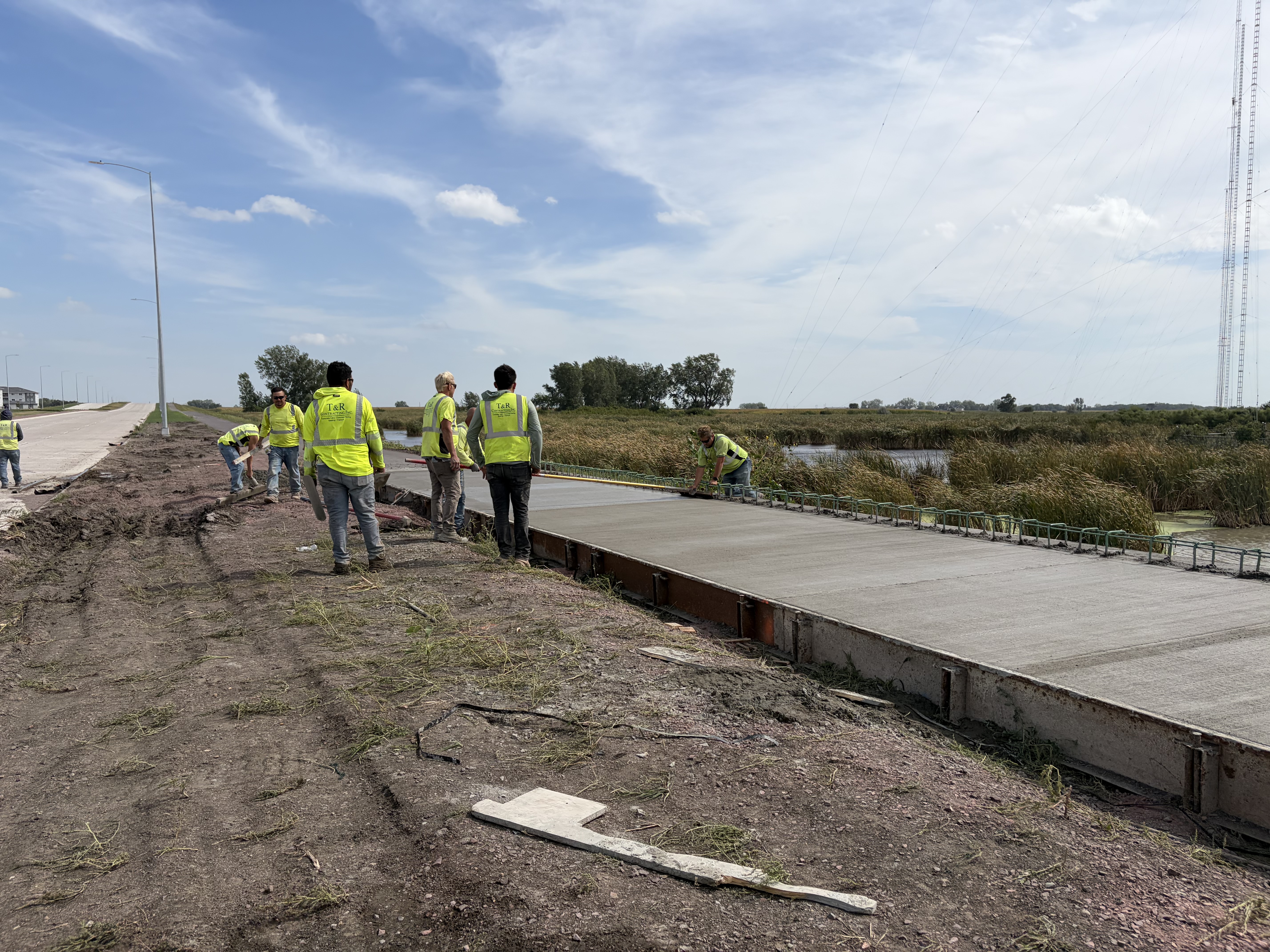 Several construction workers in high-visibility safety clothing finish and inspect a fresh concrete path alongside a wetland, with open land, utility structures, and a partly cloudy sky in the background.
