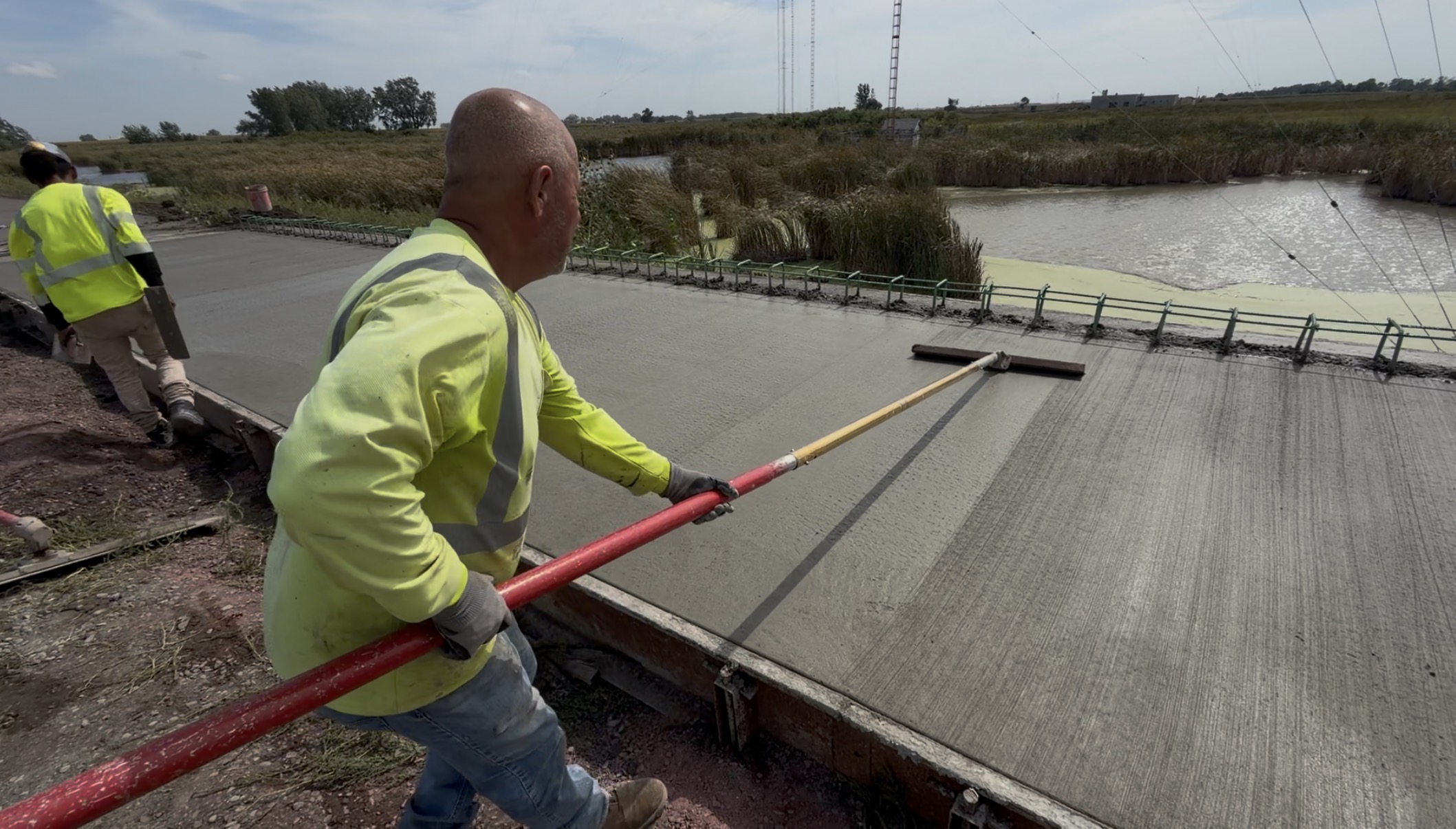 Construction worker in a high-visibility safety shirt uses a long-handled concrete finishing tool to smooth freshly poured concrete beside a wetland area with reeds and standing water.